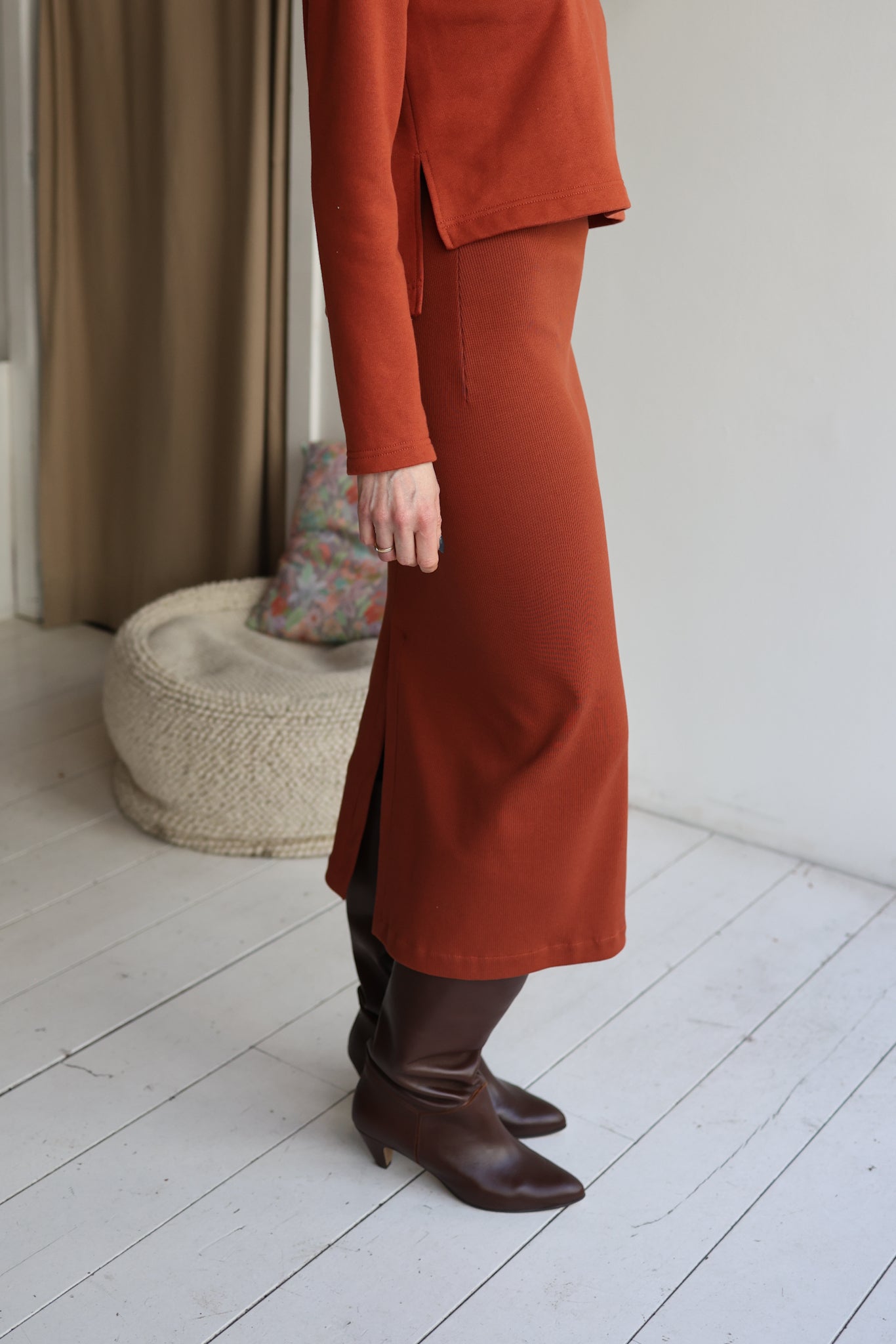 Person wearing a rust-colored suit with a skirt and brown boots in a room with a textured rug and neutral walls.