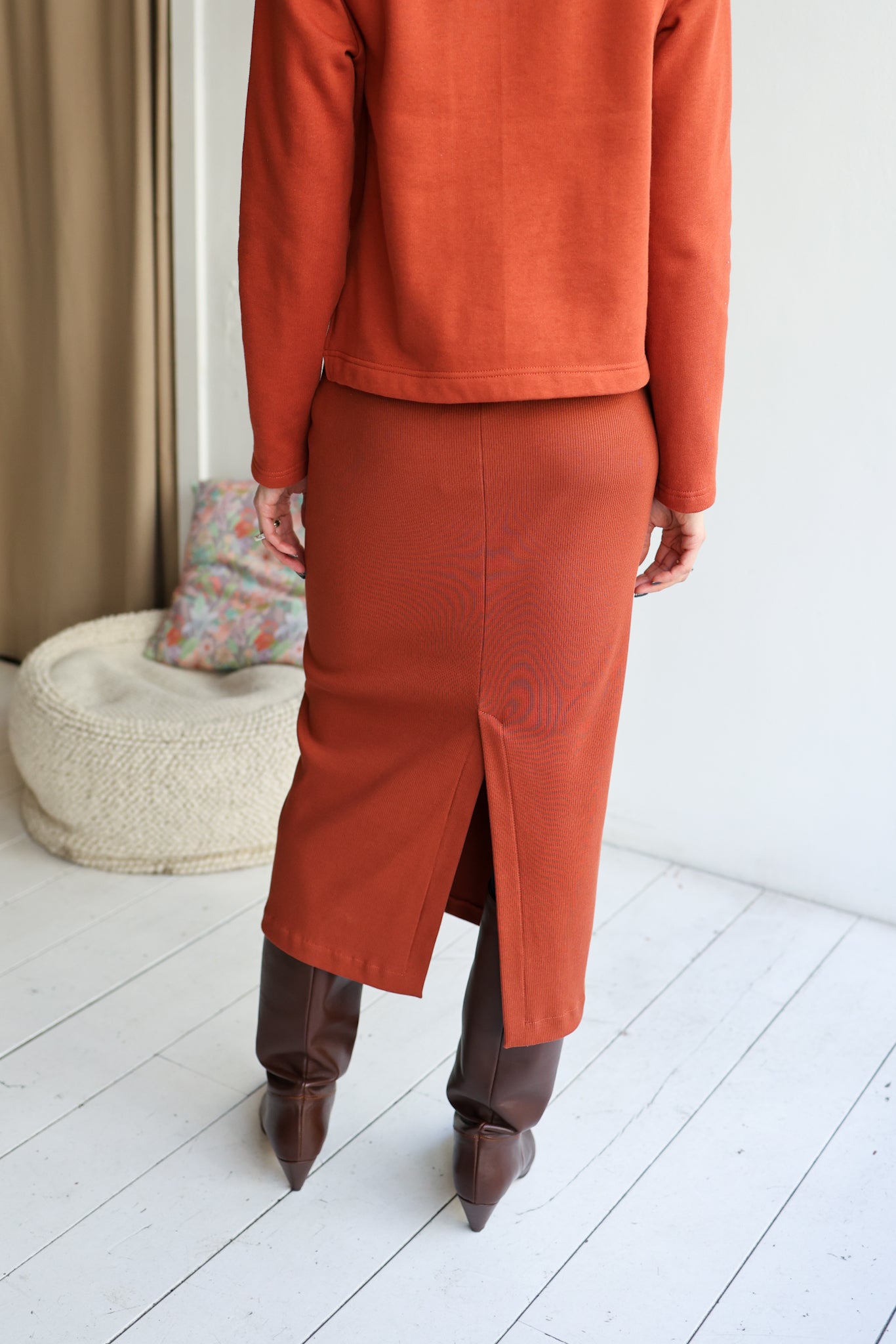 Person wearing an orange outfit with a skirt and brown boots in a minimal indoor setting.