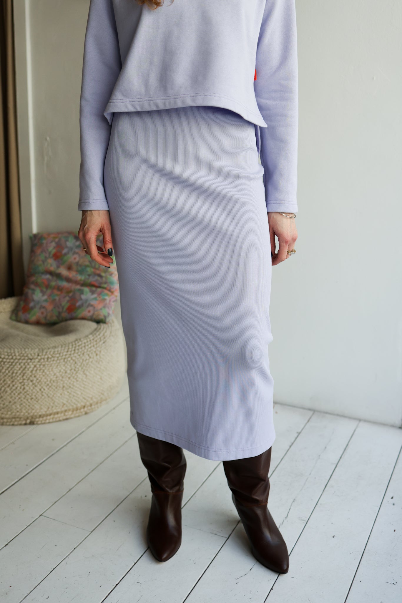 Person wearing a light purple outfit with a skirt and brown boots in a minimalistic room.