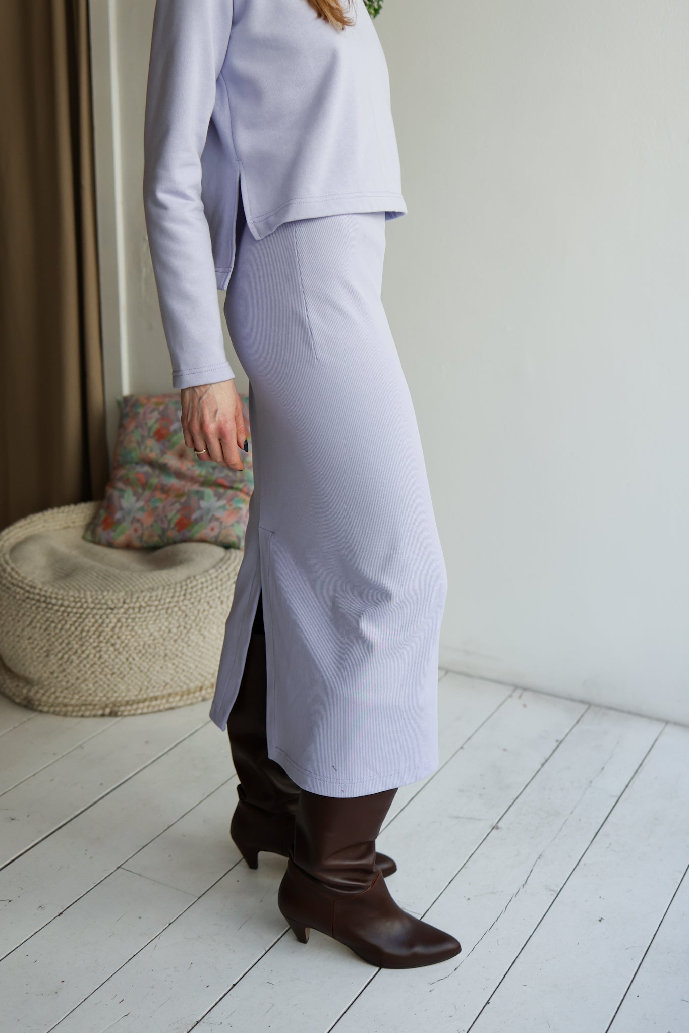 Person wearing a light purple outfit with a skirt and brown boots in a room with a white wall and wooden floor.