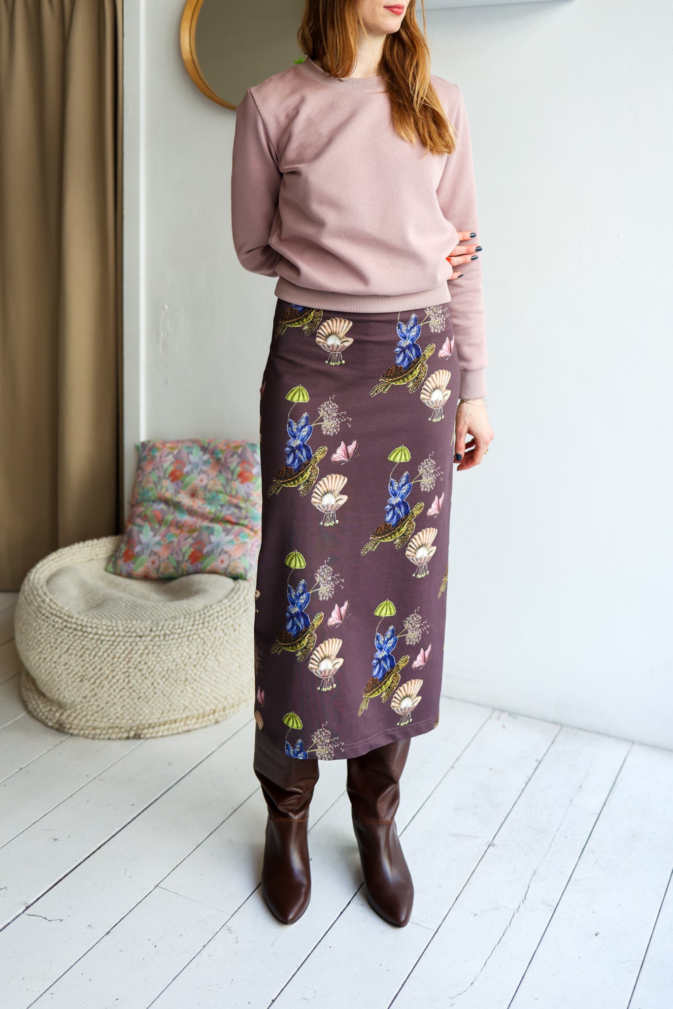 Woman wearing a pink sweater and patterned skirt standing in a room with a textured chair and mirror.