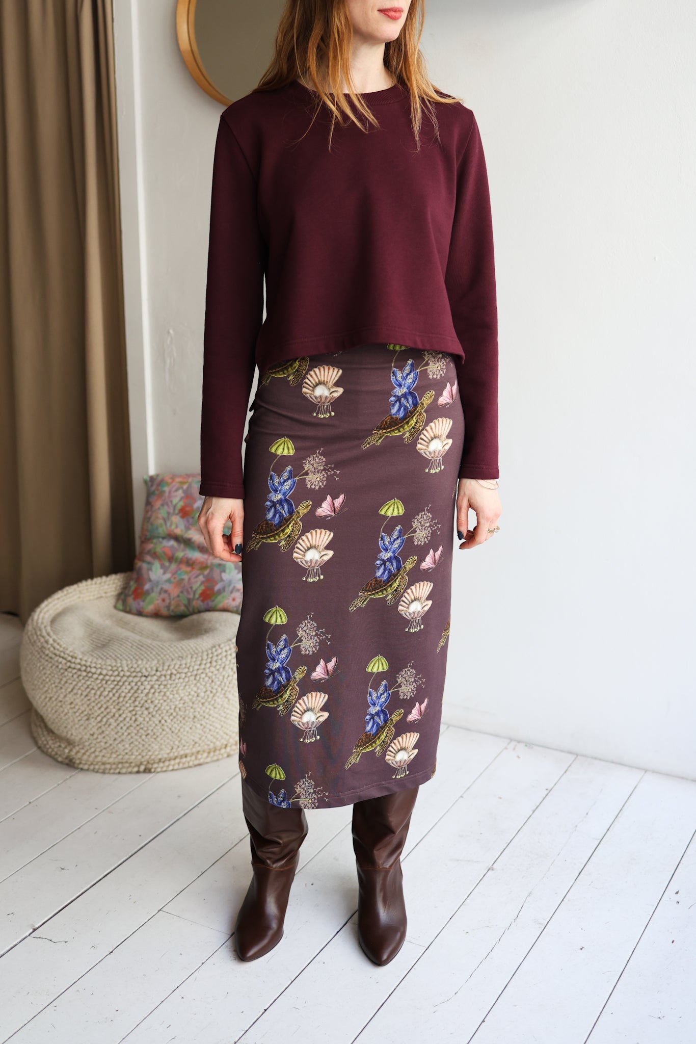 Woman wearing a burgundy sweater and patterned skirt standing in a room with a white wall and wooden floor.