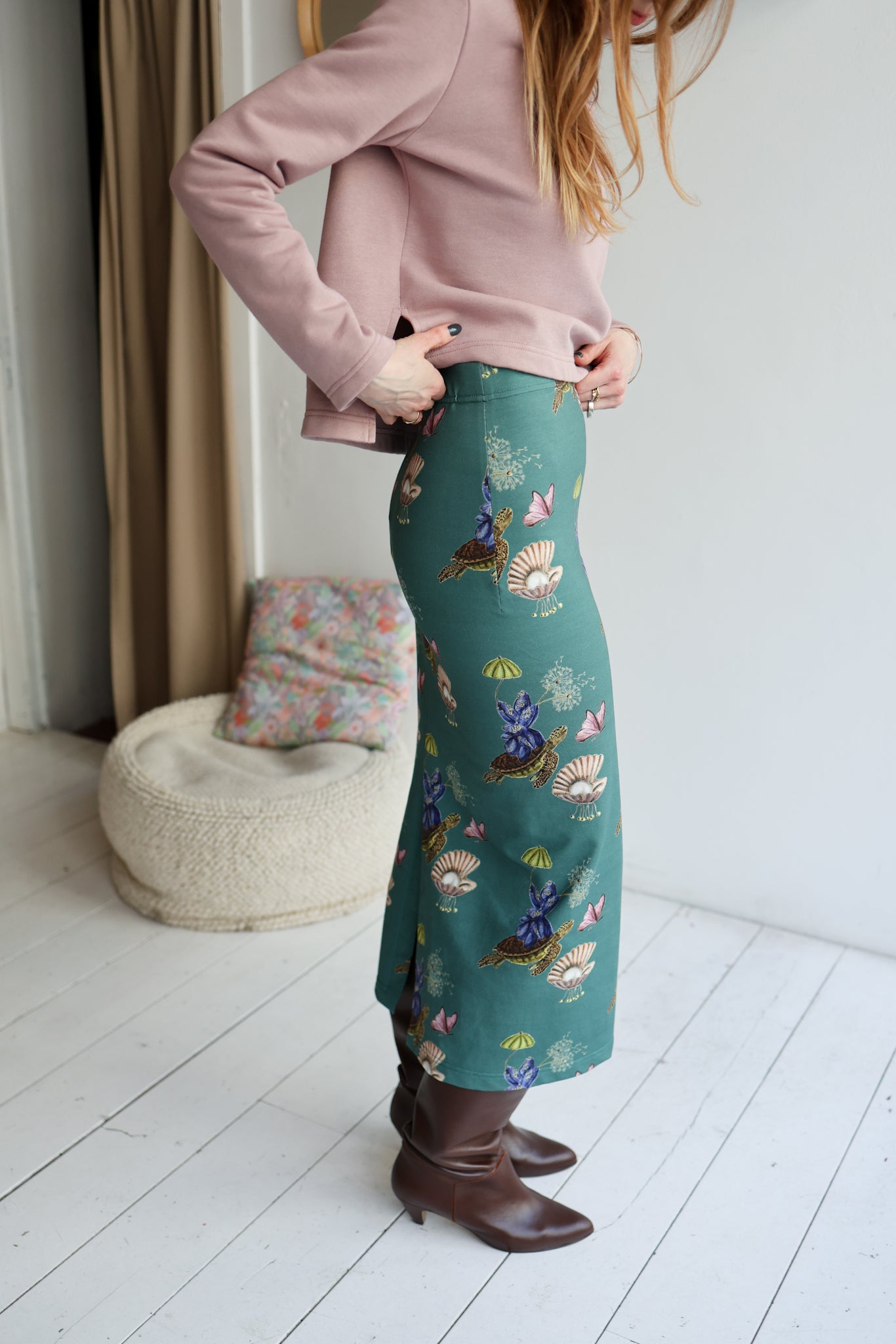 Person wearing a pink top and teal patterned skirt in a room with a white floor and beige curtain.