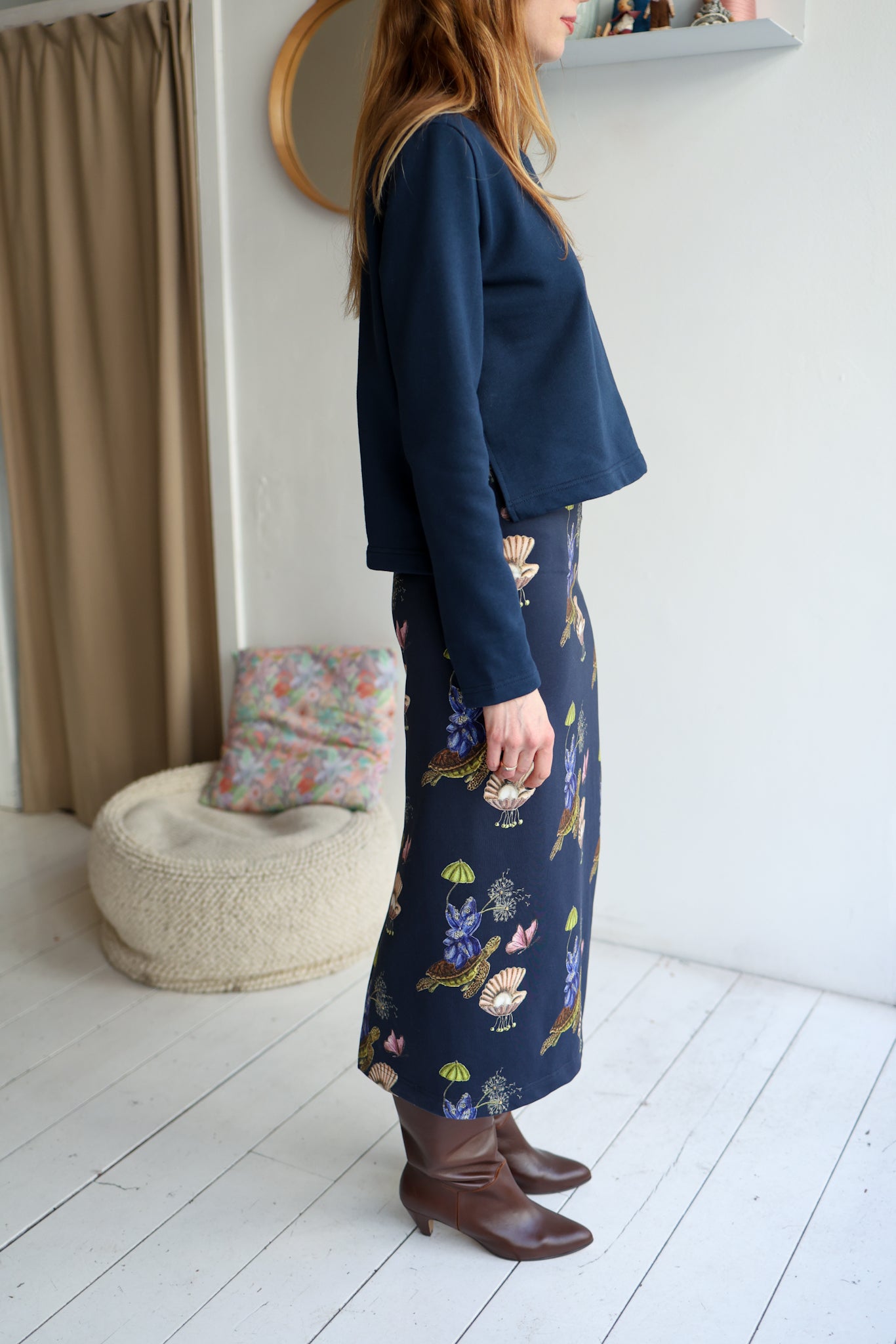 Person wearing a navy blue top and patterned skirt standing in a room with white walls and a curtain.