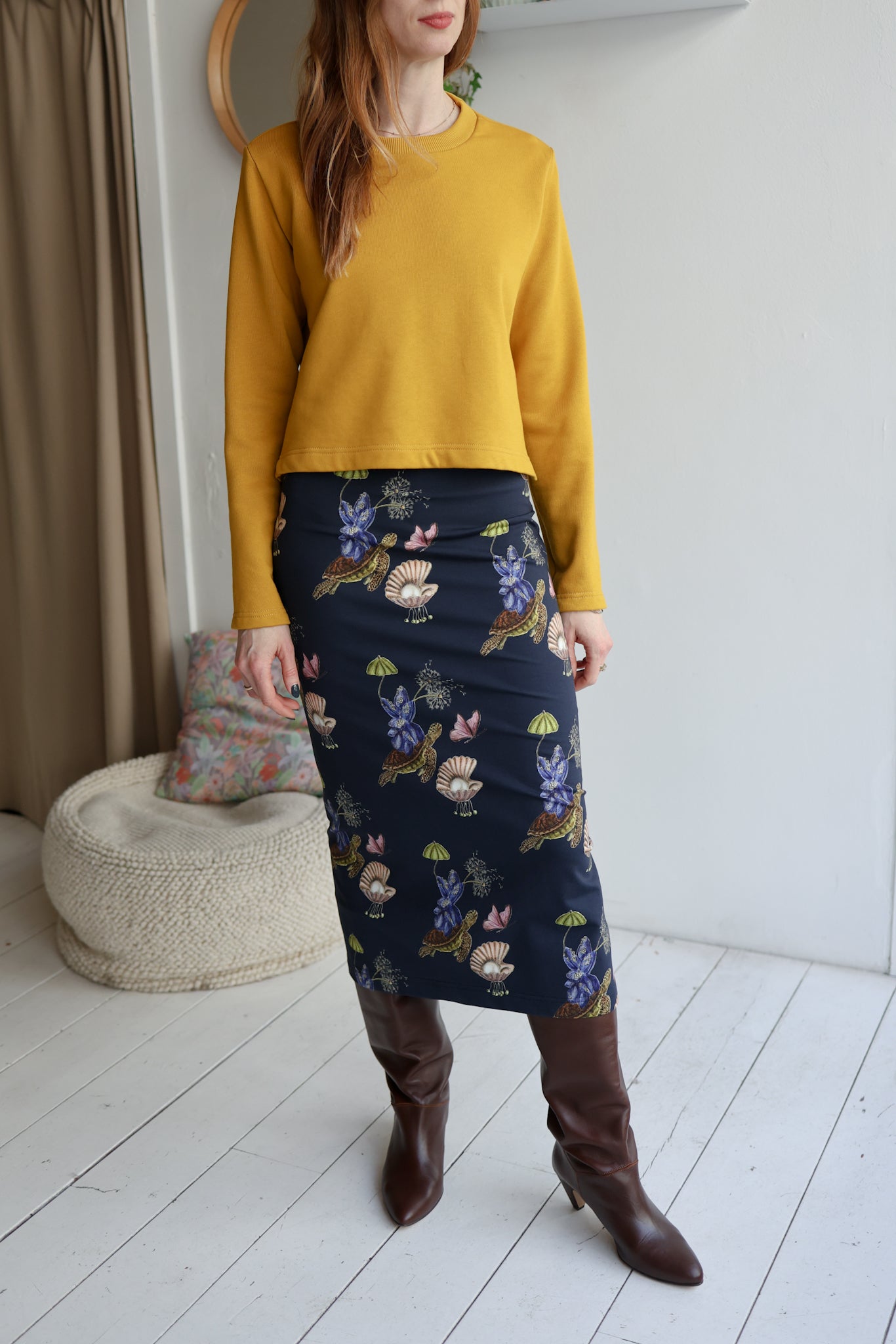 Person wearing a mustard yellow sweater and a dark blue floral skirt in a room with white walls and a wooden floor.