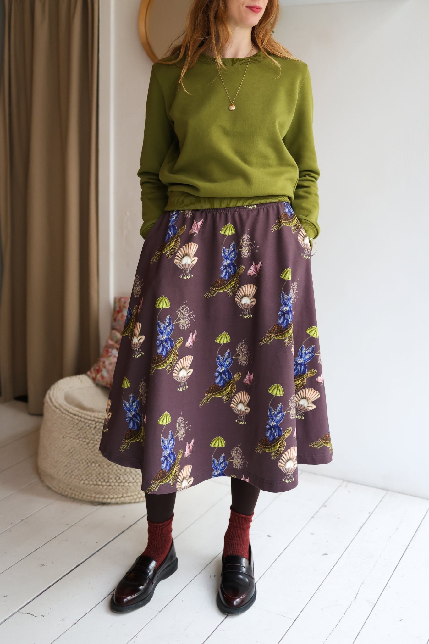 Woman wearing a green sweater and patterned skirt standing in a room with a white wall and wooden floor.