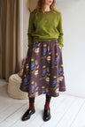Woman wearing a green sweater and patterned skirt standing in a room with a white wall and wooden floor.