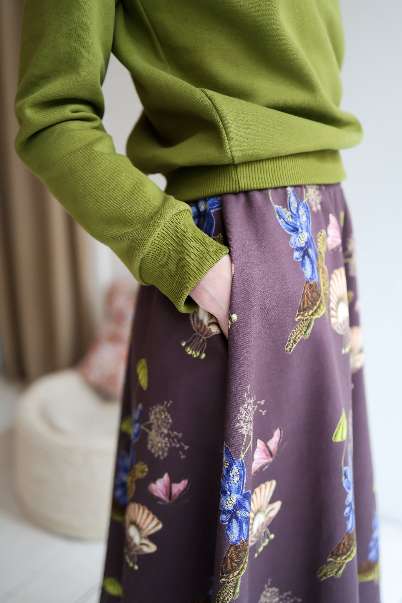 Woman wearing a green sweater and patterned skirt standing in a room with a blurred background.