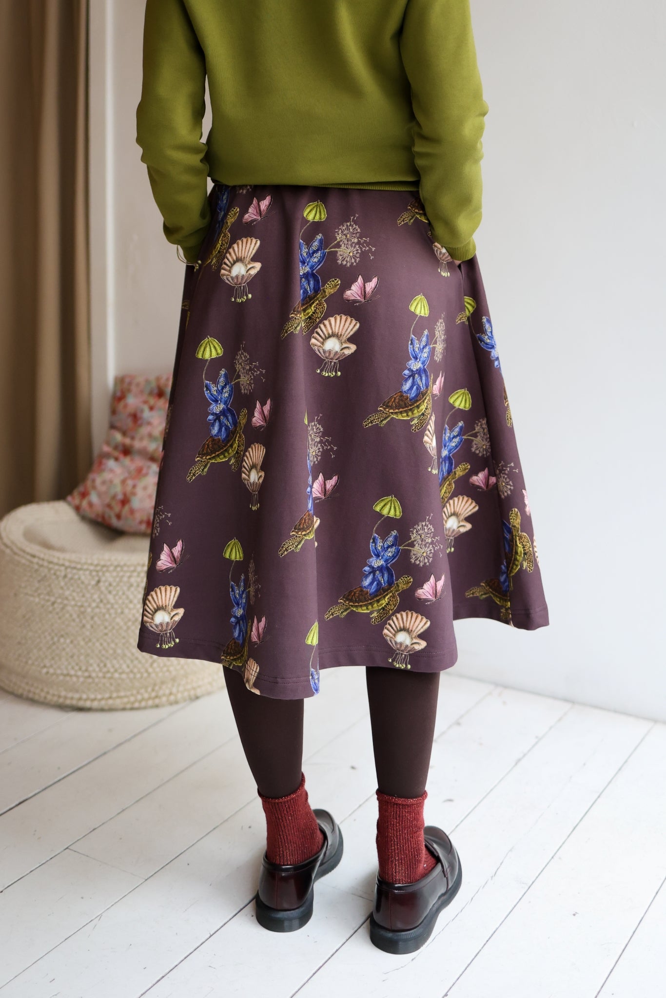 Woman wearing a green sweater and patterned skirt standing in a room with a white wall and wooden floor.
