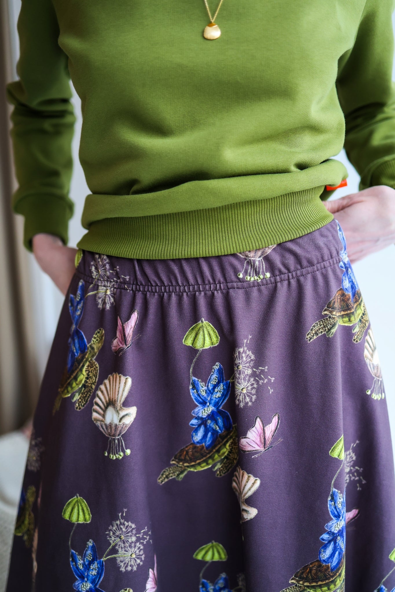 Woman wearing a green sweater and patterned skirt standing in a room with a blurred background.