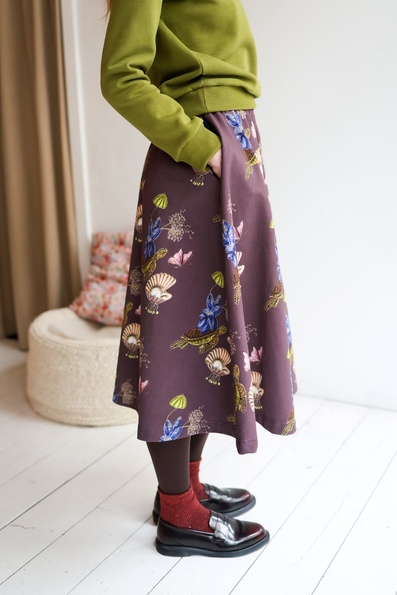 Woman wearing a green sweater and patterned skirt standing in a room with a white wall and wooden floor.