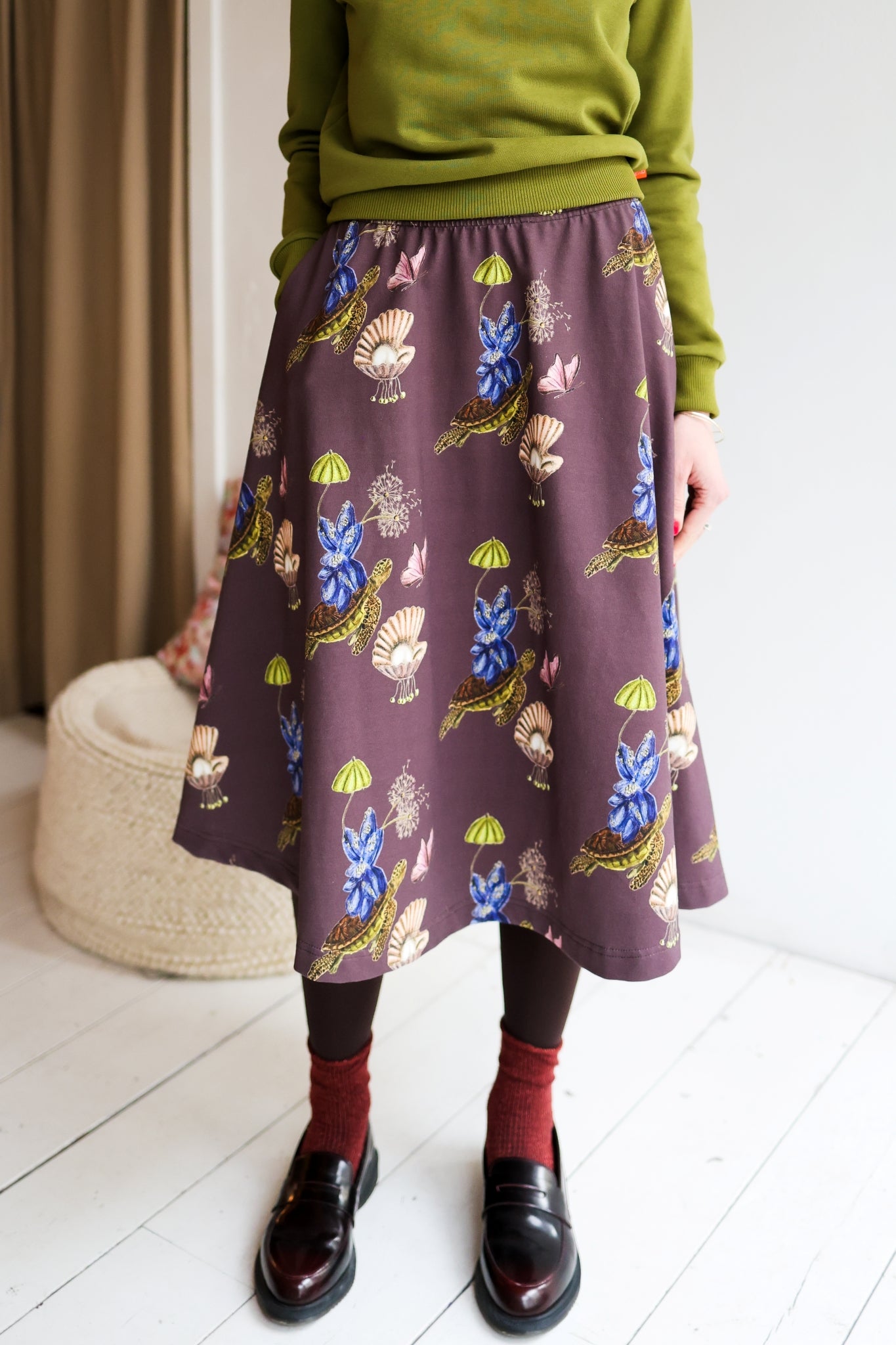 Woman wearing a green sweater and patterned skirt standing in a room with a white wall and wooden floor.