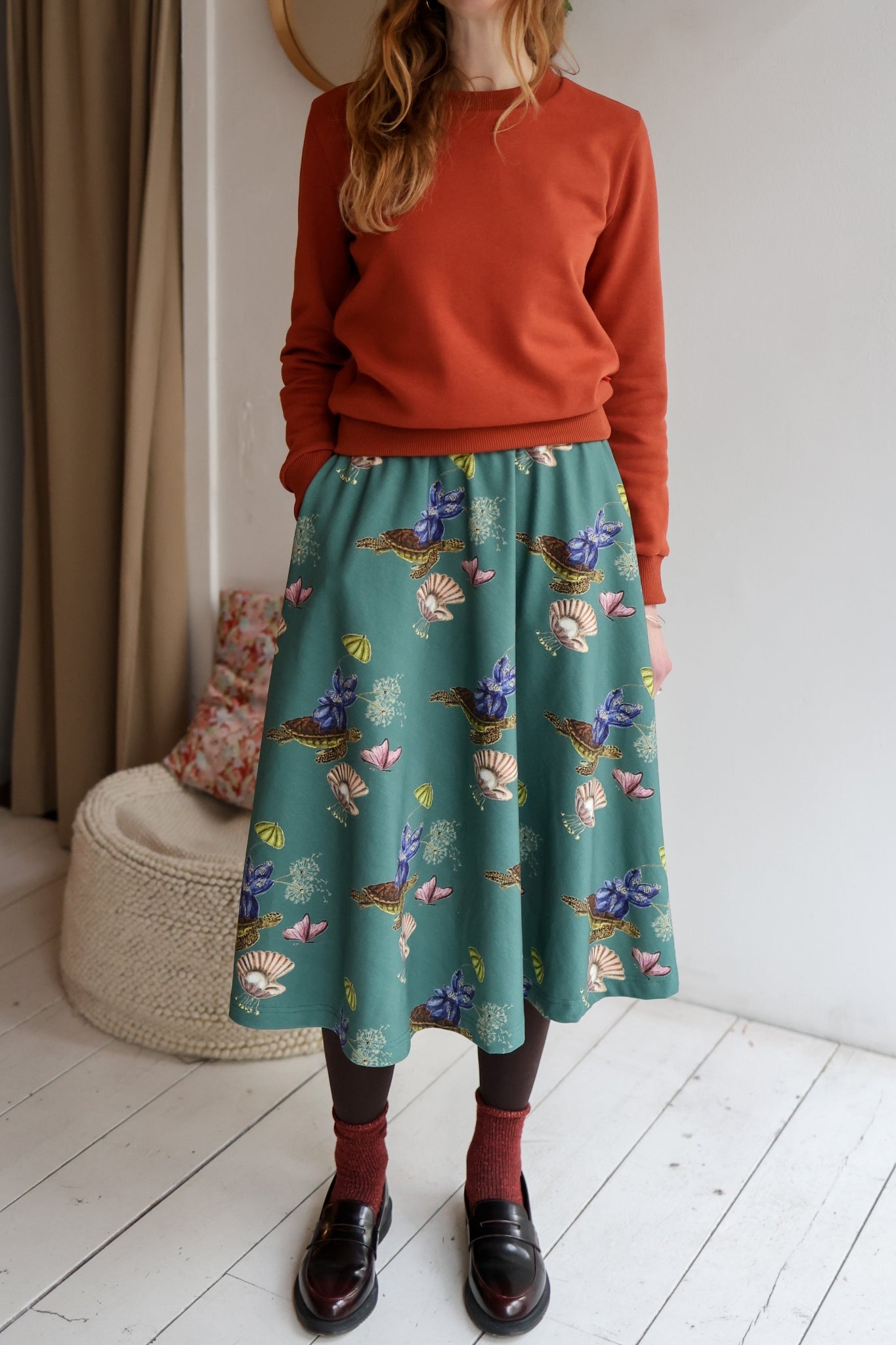 Woman wearing a red sweater and patterned skirt standing in a room with a white wall and wooden floor.