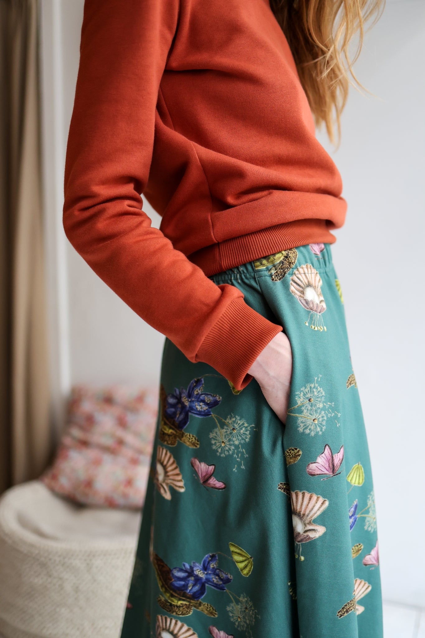 Woman wearing a red sweater and patterned skirt standing in a room with a blurred background.
