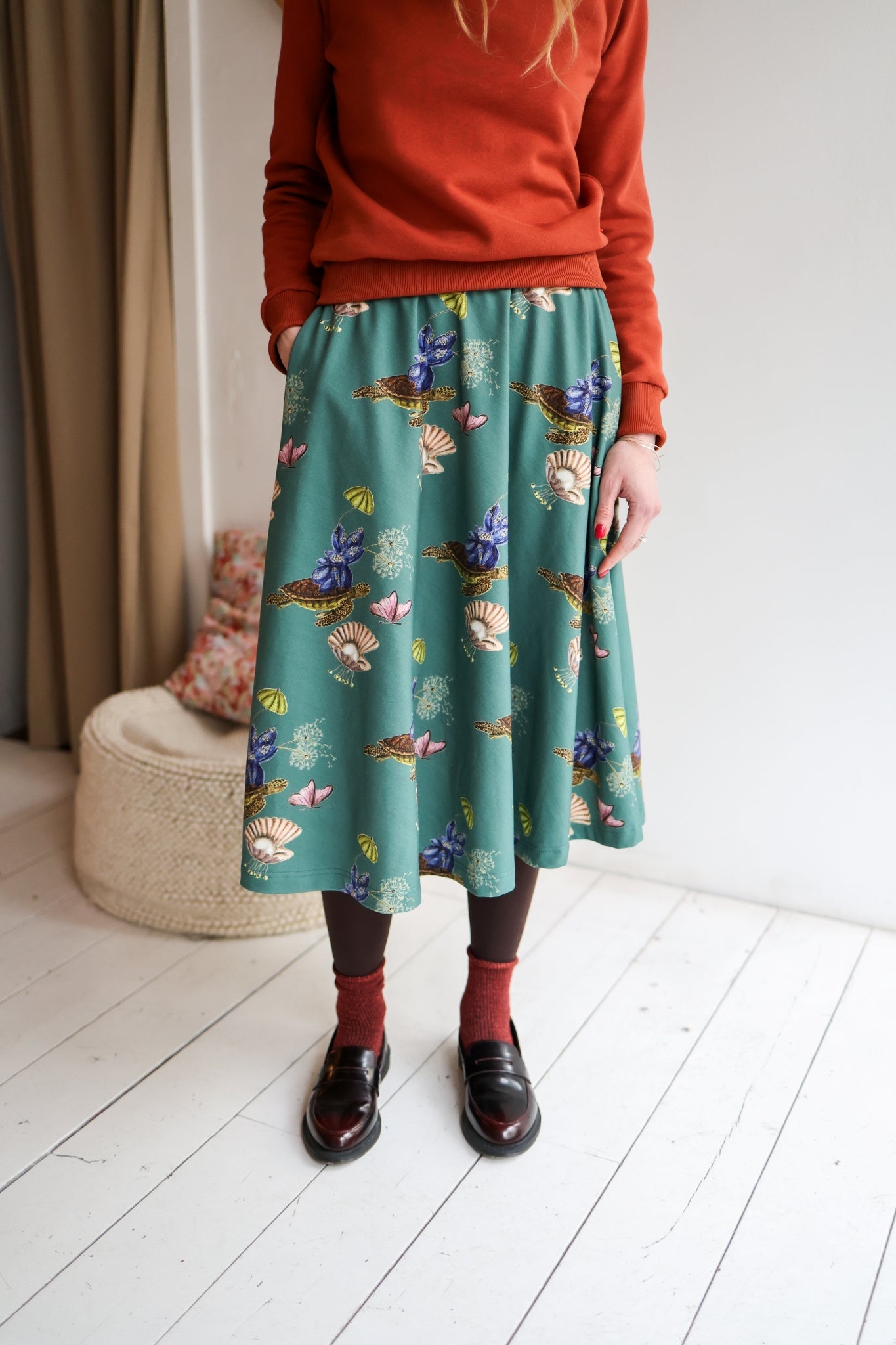 Woman wearing a red sweater and patterned skirt standing in a room with a white wall and wooden floor.