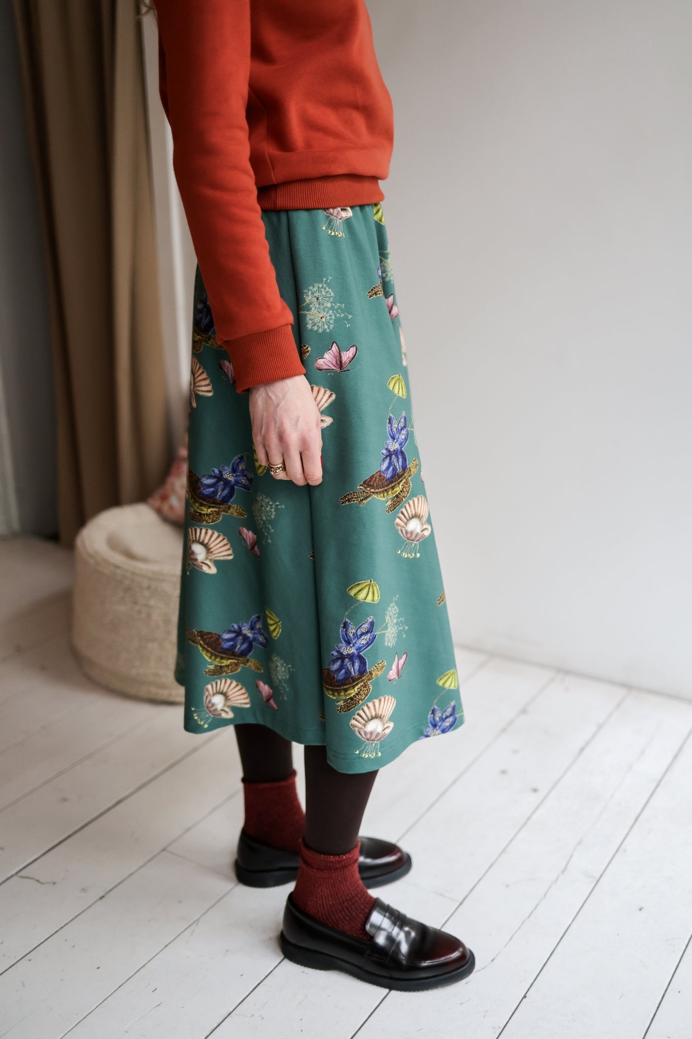 Woman wearing a red sweater and patterned skirt standing in a room with a white wall and wooden floor.