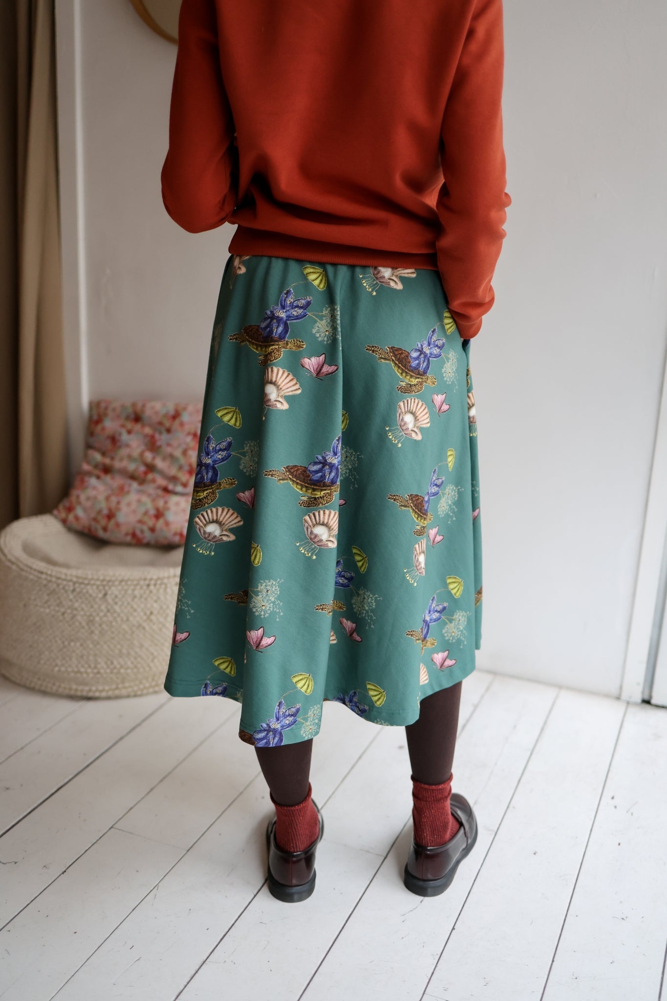 Woman wearing a red sweater and patterned skirt standing in a room with a white wall and wooden floor.