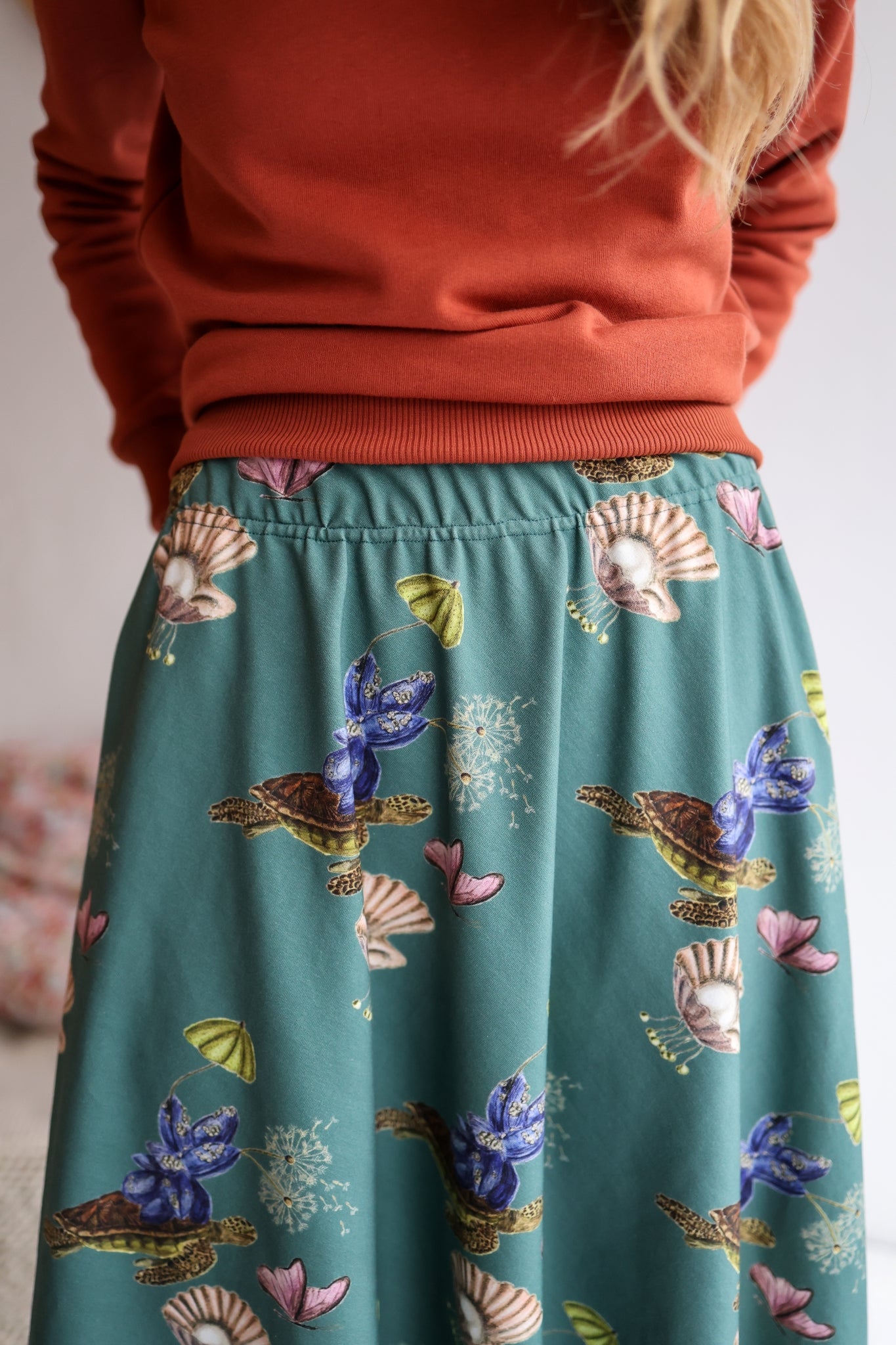 Woman wearing a red sweater and patterned skirt standing in a room with a blurred background.