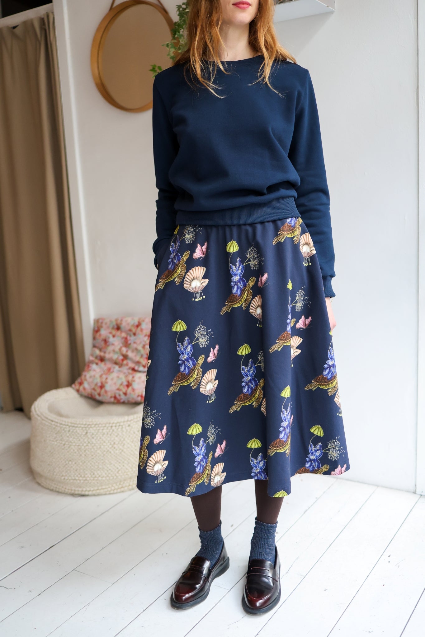 Woman wearing a navy sweater and patterned skirt in a room with a white wall and wooden floor.
