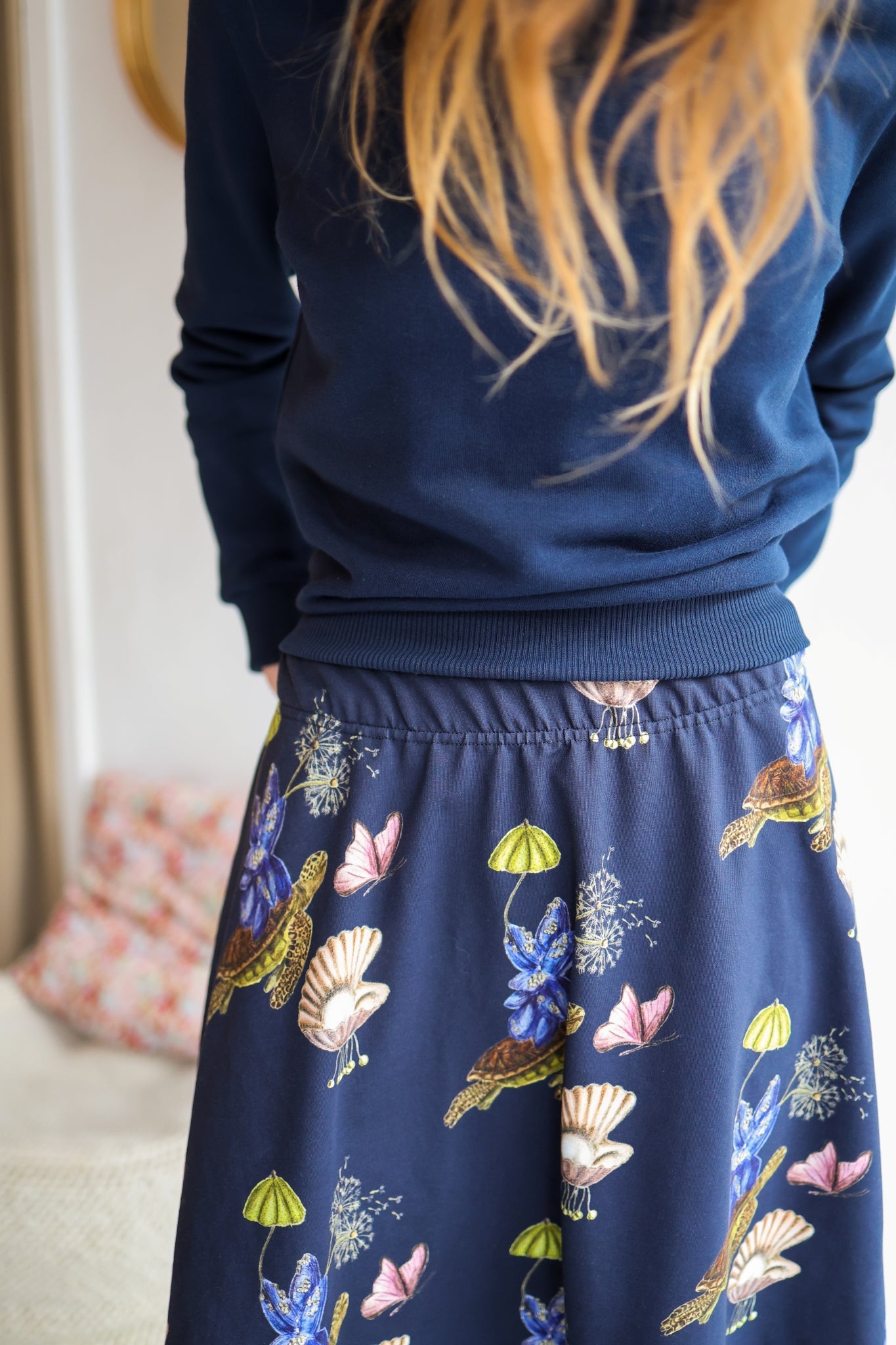 Close-up of a woman wearing a navy sweater and patterned skirt in a room with a blurred background.