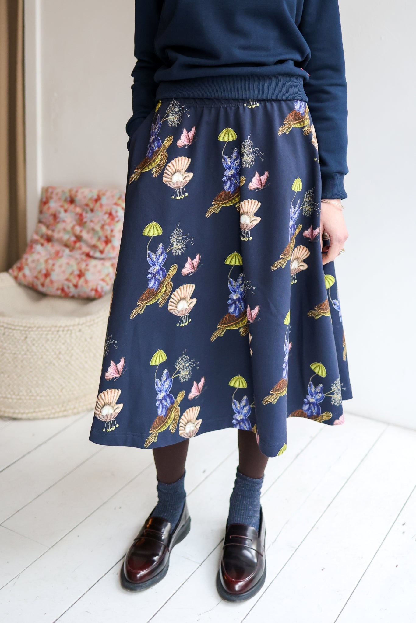 Woman wearing a navy sweater and patterned skirt in a room with a white wall and wooden floor.