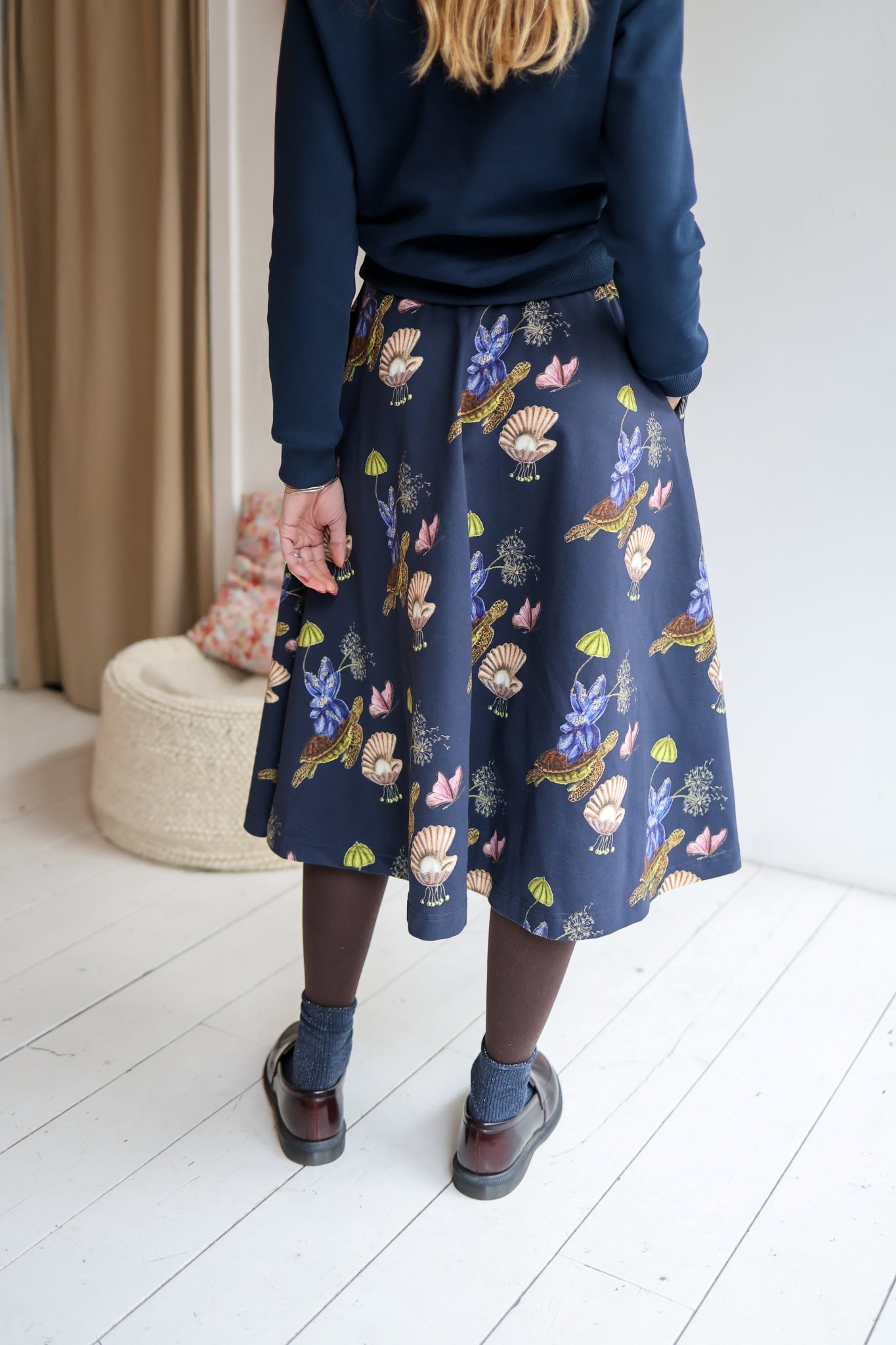 Woman wearing a navy sweater and patterned skirt in a room with a white wall and wooden floor.
