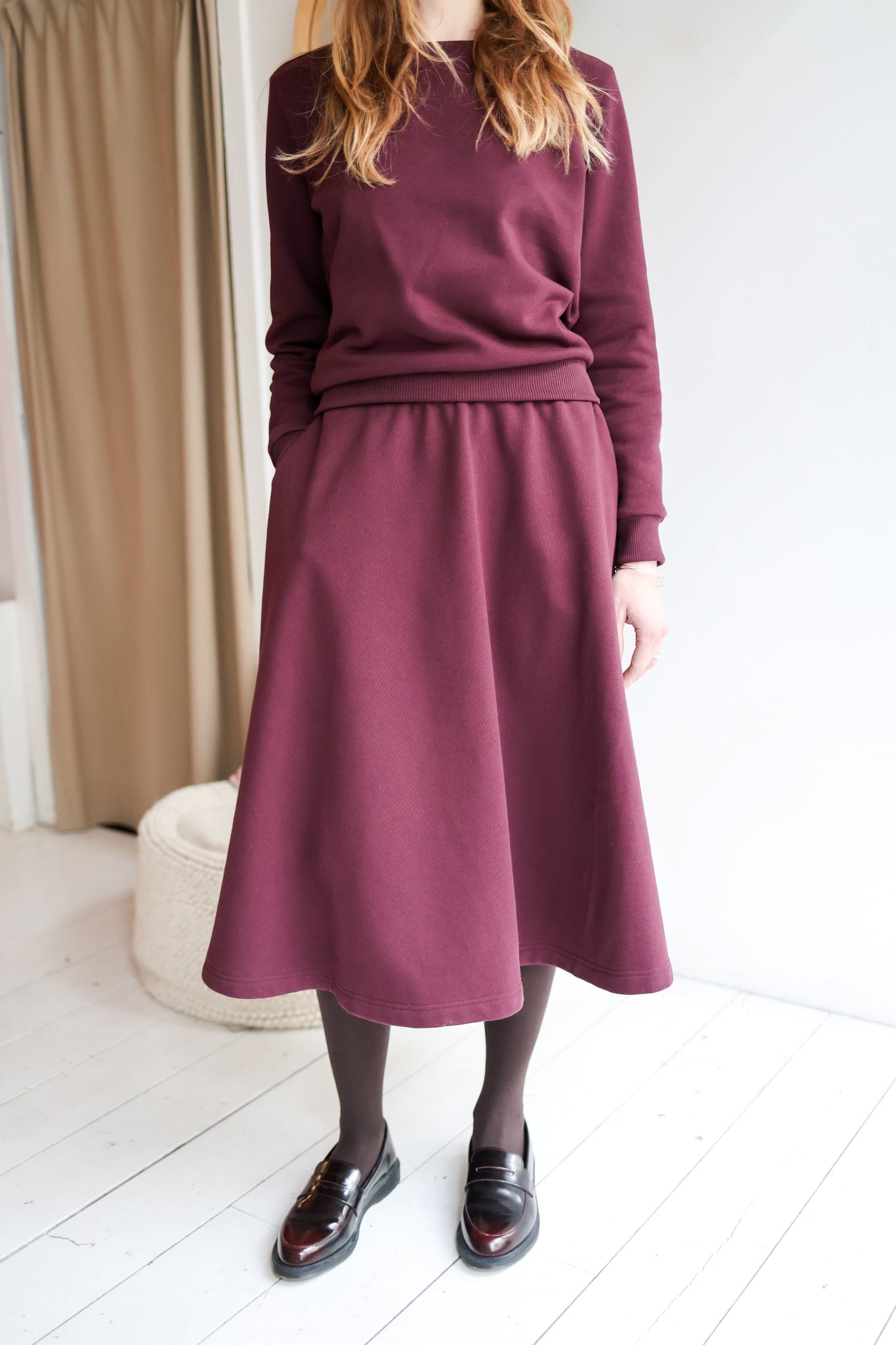 Woman wearing a bordeaux outfit with a skirt and sweater in a minimal indoor setting.