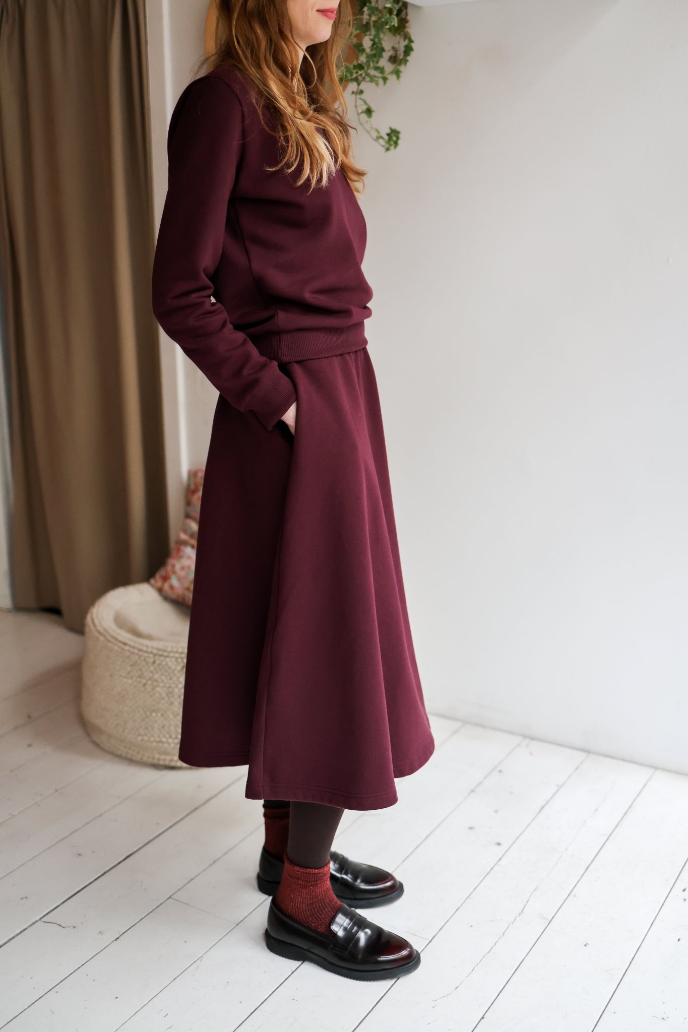 Woman wearing a bordeaux outfit with a skirt and sweater in a minimal indoor setting.