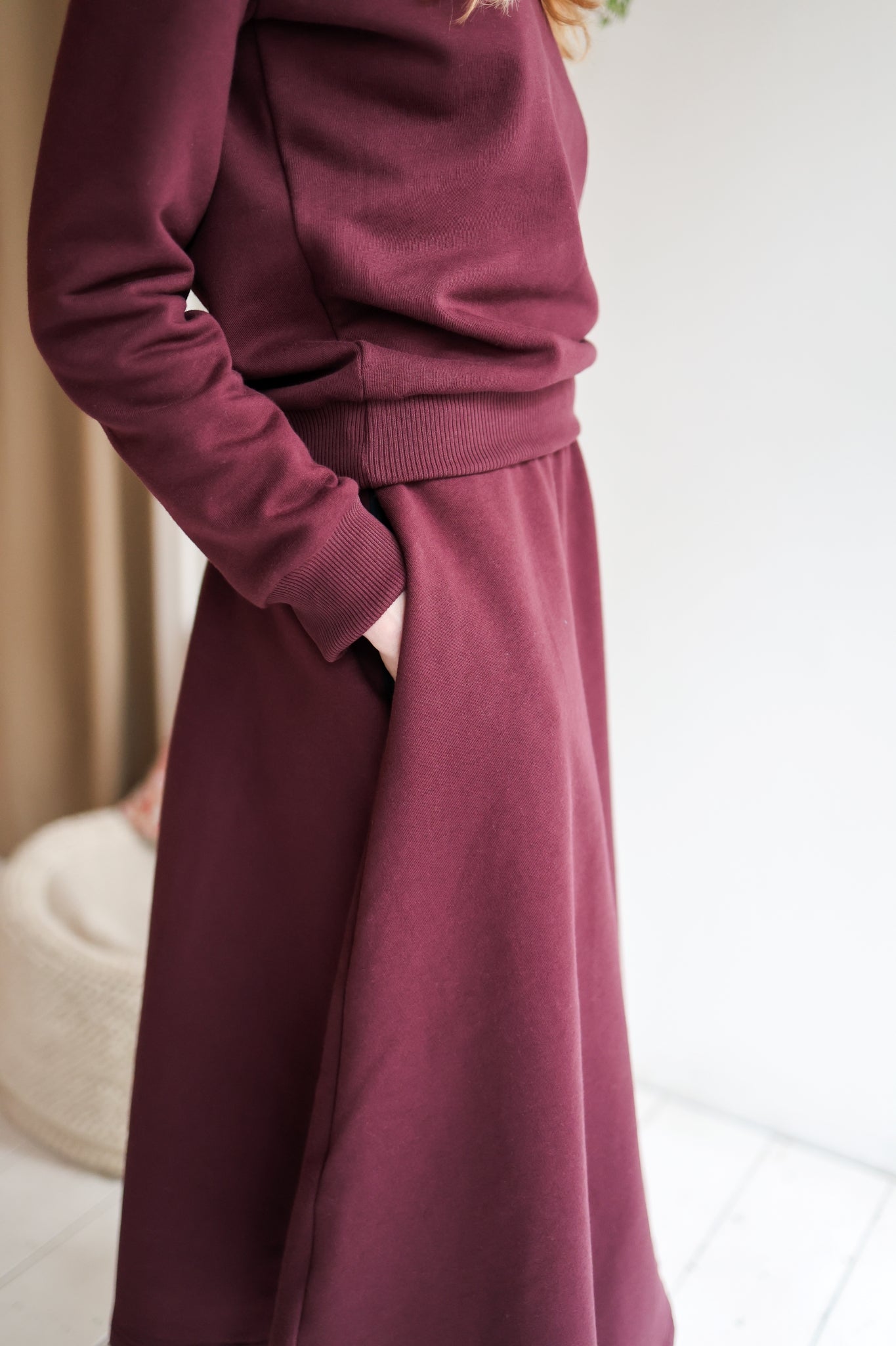 Woman wearing a bordeaux outfit with a skirt and sweater in a minimal indoor setting.