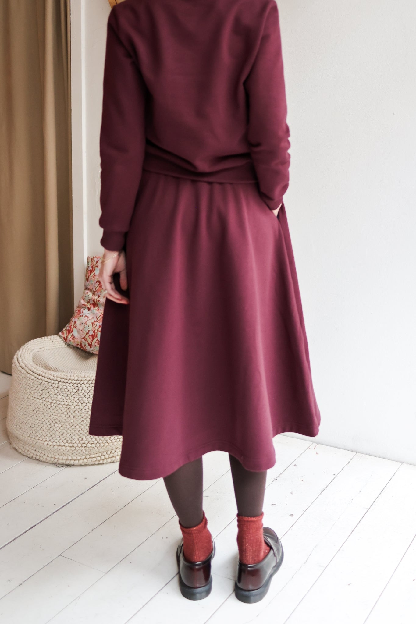 Woman wearing a bordeaux outfit with a skirt and sweater in a minimal indoor setting.