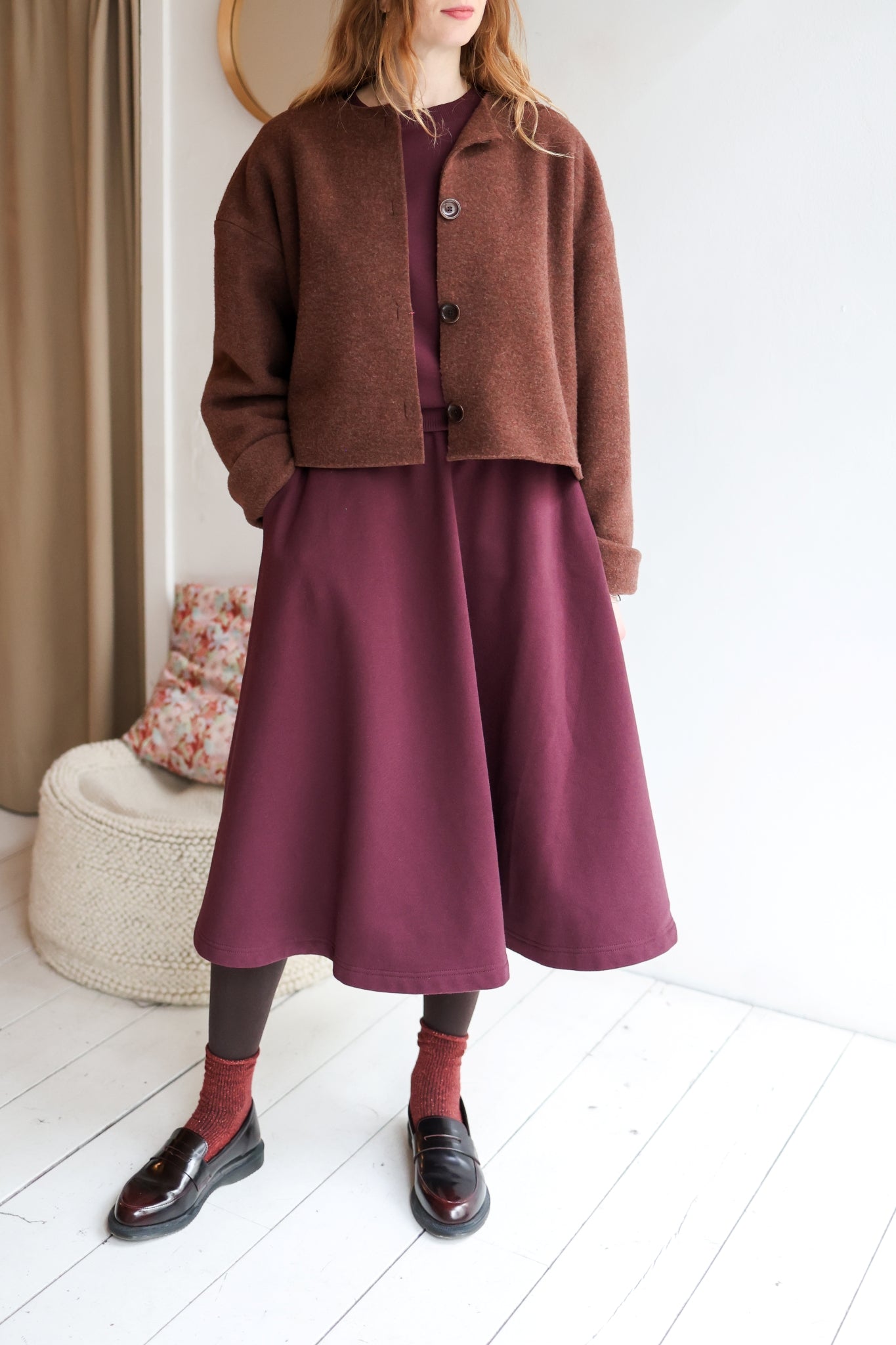 Woman wearing a brown coat with a bordeaux outfit in a minimal indoor setting.
