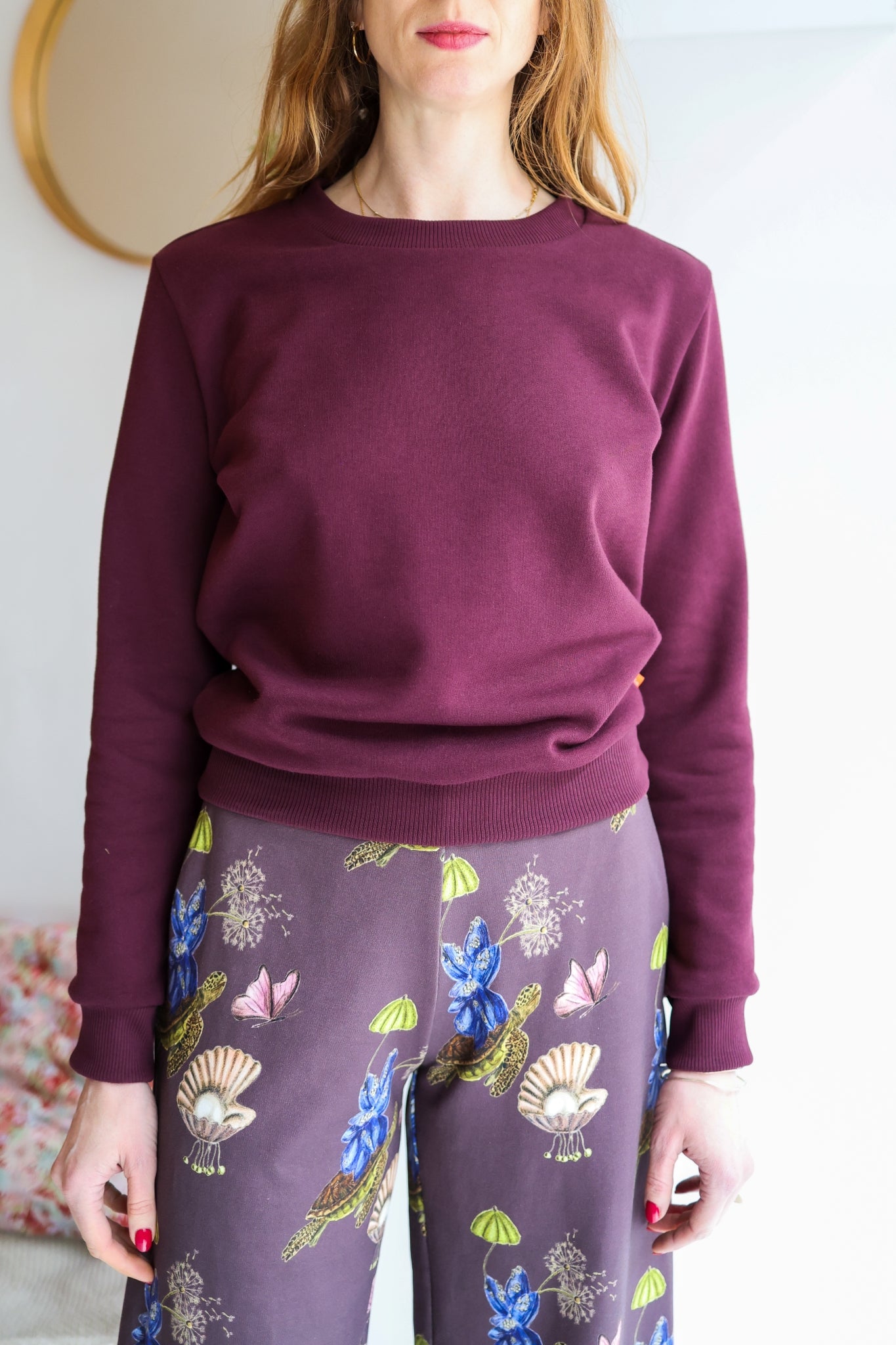 Person wearing a bordeaux sweater and patterned pants against a white wall.