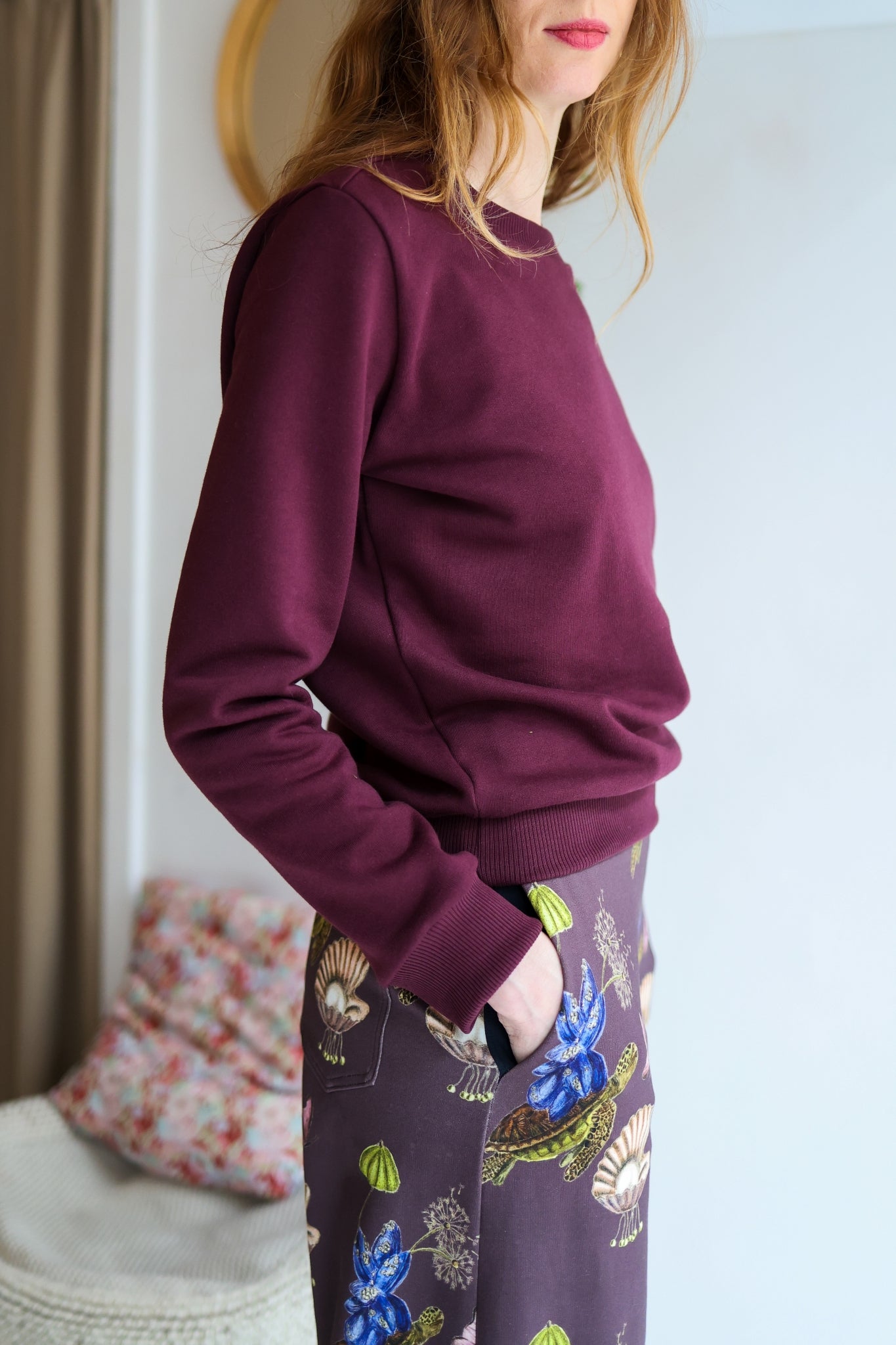 Person wearing a bordeaux sweatshirt and patterned pants in a room with a floral pillow and white wall.
