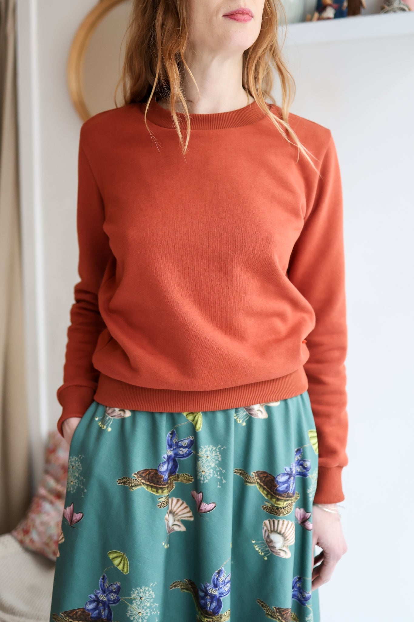 Woman wearing a rust-colored sweatshirt and a floral green skirt indoors.