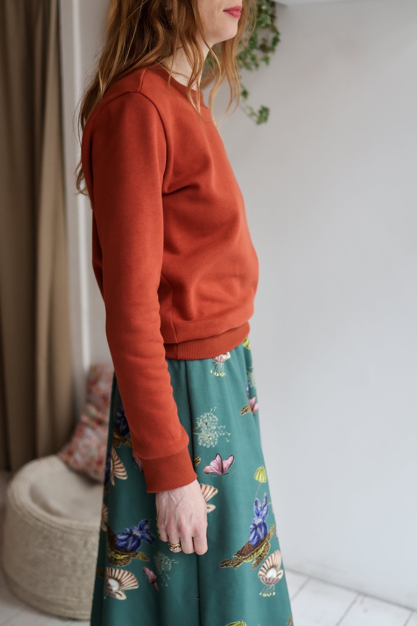 Person wearing a rust-colored sweatshirt and a floral green skirt indoors.