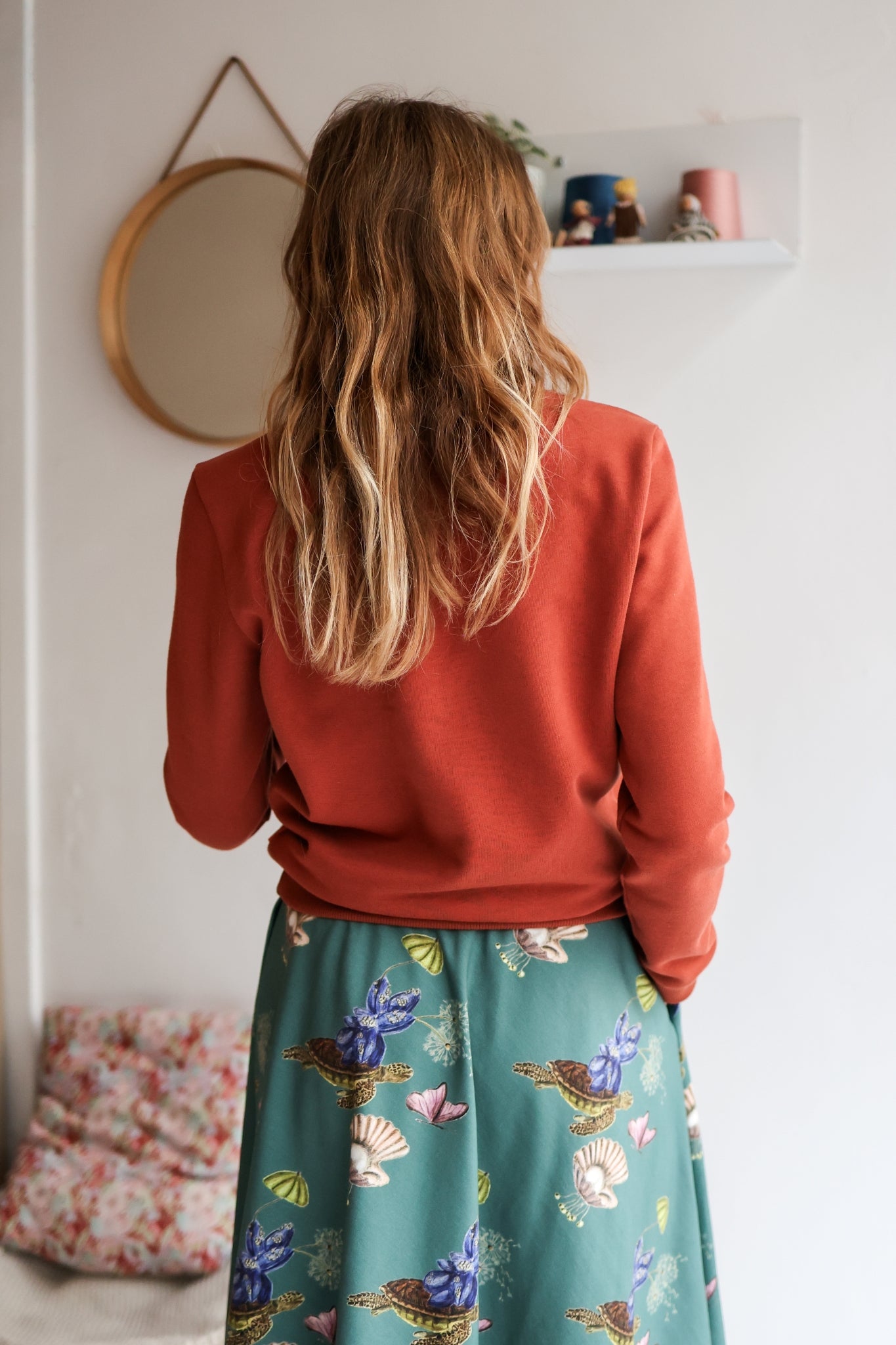 Person wearing a rust-colored sweater and teal floral skirt standing in a room with a mirror and shelf.