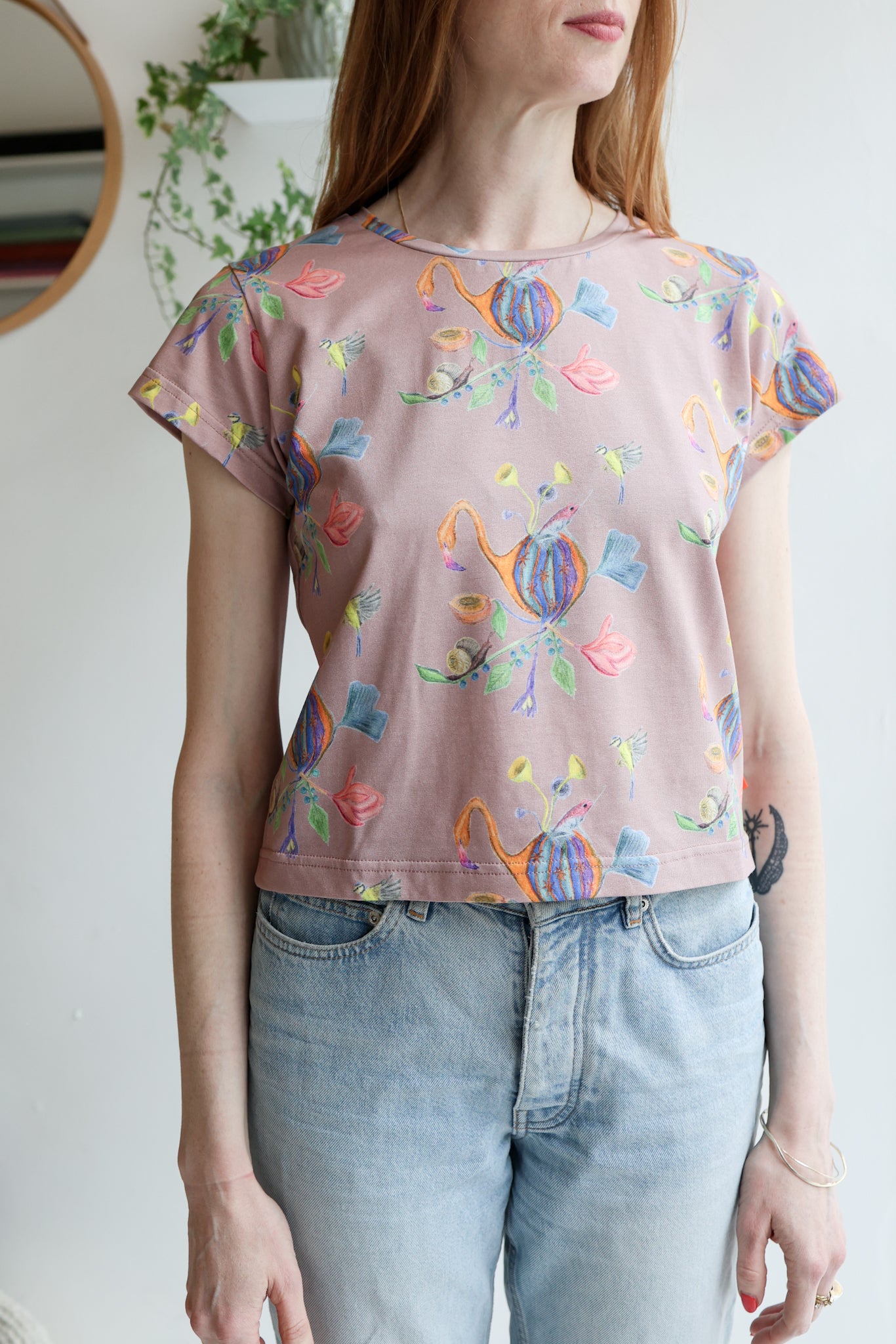 Woman wearing a pink t-shirt with colorful patterns and light blue jeans indoors.