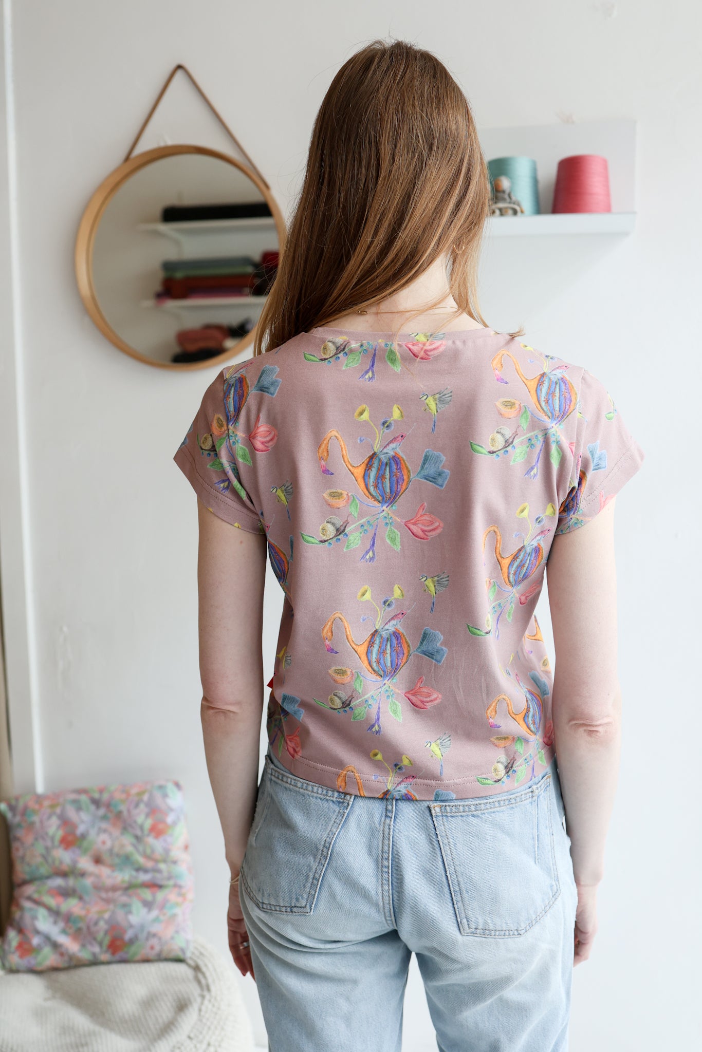 Woman wearing a pink t-shirt with colorful patterns and light blue jeans indoors.