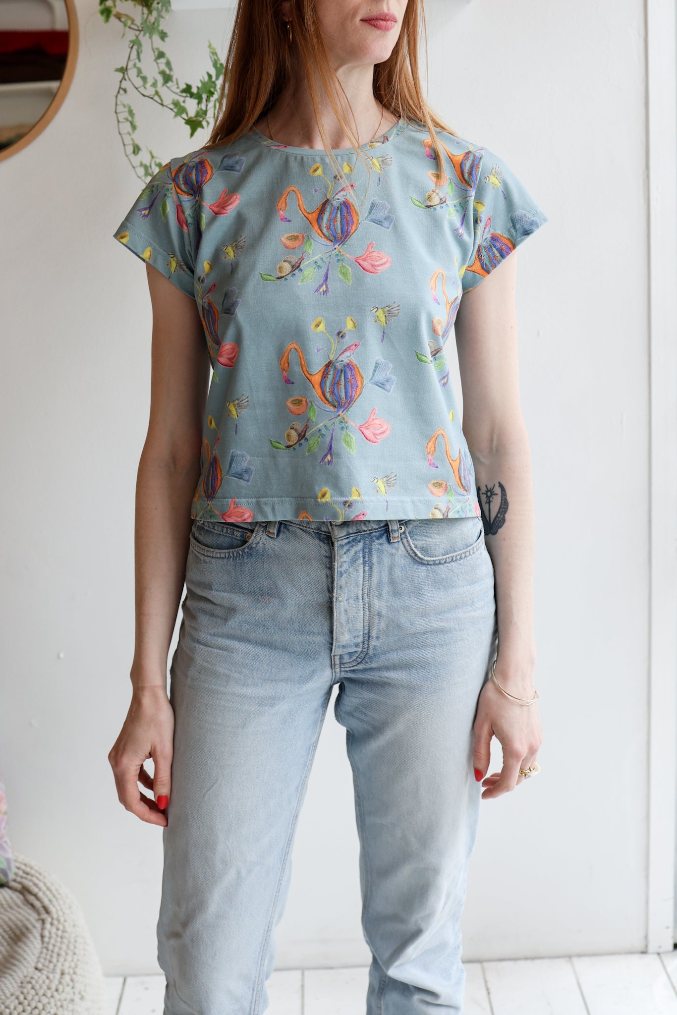 Woman wearing a blue t-shirt with colorful patterns and light blue jeans indoors.