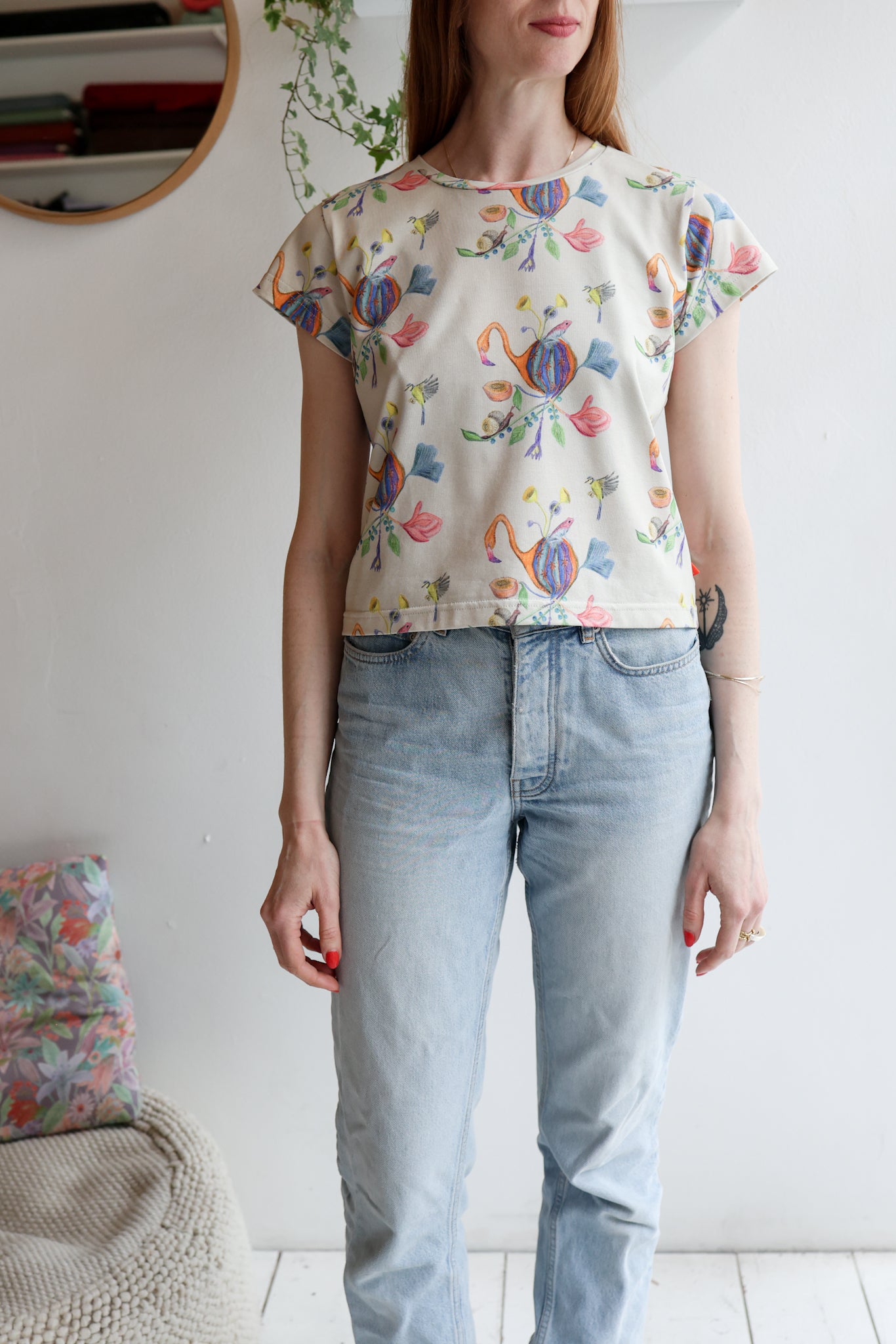 Woman wearing a beige t-shirt with colorful patterns and light blue jeans indoors.